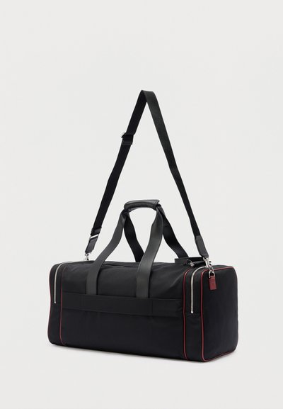 Μαύρη ορθογώνια σακούλα τύπου duffel με διπλές χειρολαβές, ρυθμιζόμενο λουράκι ώμου, ασημένια φερμουάρ και διακριτική κόκκινη επένδυση σε απλή φόντο.