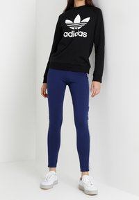 Felpa nera con logo Adidas bianco, abbinata a leggins blu con strisce laterali bianche e scarpe da ginnastica bianche. Texture liscia.