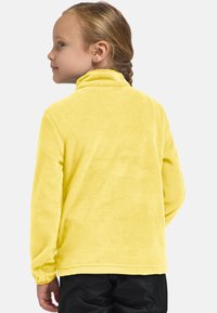 Kind met gevlochten haar, gekleed in een heldere gele fleecjas en zwarte broek, zijwaarts gericht met een lichte glimlach.