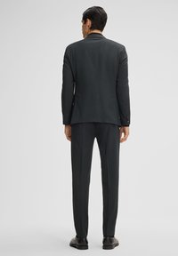 Donkergrijs pak met een slim fit, met een gestructureerd patroon. Het jasje heeft een notch-kraag, twee achterventilaties en op maat gemaakte broeken. Zwarte schoenen.