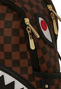 Zaino a scacchi marrone con accenti neri, caratterizzato da un design dell'occhio rosso e bianco molto evidente e hardware in metallo dorato. Tessuto testurizzato.