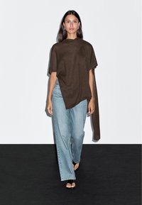 Kobieta idąca po czarnym podłożu, ubrana w brązowy asymetryczny ponczo, jasnoniebieskie szerokie dżinsy, czarne sandały na obcasie oraz srebrne kolczyki.