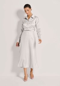 Woman wearing a light gray satin long-sleeve shirt and matching midi skirt, paired with pink pointed-toe heels, standing against plain wall.