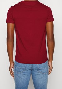 Bordeauxfarbene Baumwoll-T-Shirt mit kurzen Ärmeln und Rundhalsausschnitt, getragen mit blauen Jeans aus Denim, die über doppelte Nähte und Gesäßtaschen verfügen.