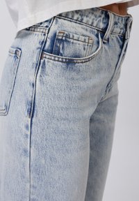 Des jeans en denim bleu clair présentent une taille haute, un design à cinq poches, des accents de couture en orange et une texture délavée sur le tissu.