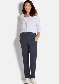 Navy textuurbroek met voorzakken en plooien, gecombineerd met een wit long-sleeve shirt. Het model draagt witte sneakers.