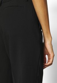 Svarta byxor med ett slätt, lätt tyg. Har sidofickor, en skräddarsydd passform och subtila sömnadsdetaljer längs sömmarna.