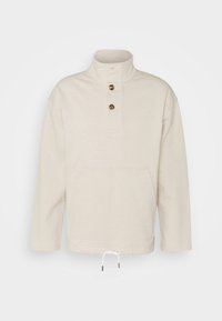 Pull beige clair avec col haut, deux boutons, poche avant kangourou et cordon blanc à l'ourlet.