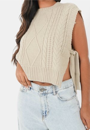 Kobieta w beżowym swetrze bez rękawów z warkoczowym splotem i wiązaniem z boku, w połączeniu z jasnoniebieskimi wysokim stanem jeansami skróconymi do talii.