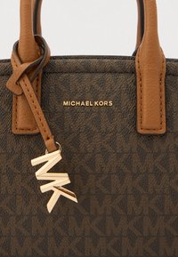 Borsa marrone con motivo a fantasia, dotata di manici in pelle color cuoio, logo dorato e un ciondolo dorato con le lettere "MK". Il materiale testurizzato aggiunge dettaglio.