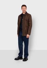 Chaqueta de cuero marrón con cierre de botones, dos bolsillos frontales y cuello. Llevada sobre una camisa negra y combinada con jeans azules y zapatos negros.