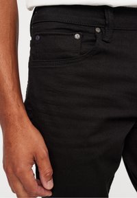 Svarta denimjeans med en åtsittande design, synliga sömmar och en framficka med metallhardware. Handen vilar på midjan.