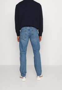 Persona de pie de espaldas, vistiendo jeans azul claro, un suéter oscuro de color navy y zapatillas blancas con rayas rojas y navy.