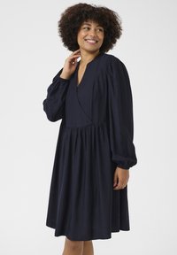 Vestido azul marino hecho de tela ligera, con escote en v, mangas largas abullonadas y una longitud por encima de la rodilla con cintura fruncida.
