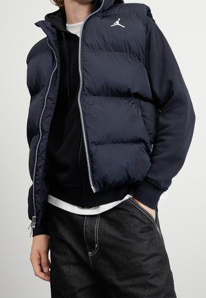 Osoba ubrana w granatową pikowaną kamizelkę z zamkiem błyskawicznym i białym logo Jordana na czarnej bluzie z kapturem oraz ciemnych dżinsach, ręka w kieszeni, neutralne tło.