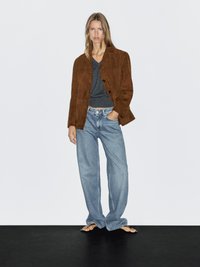 Vrouw in een bruin suède jack, donkergrijze V-hals top, losse lichtblauwe jeans en zwarte open teen sandalen, staand tegen een witte achtergrond.
