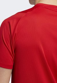 Gros plan d'une personne portant un t-shirt rouge uni à manches courtes, montrant le dos et la zone de l'épaule.