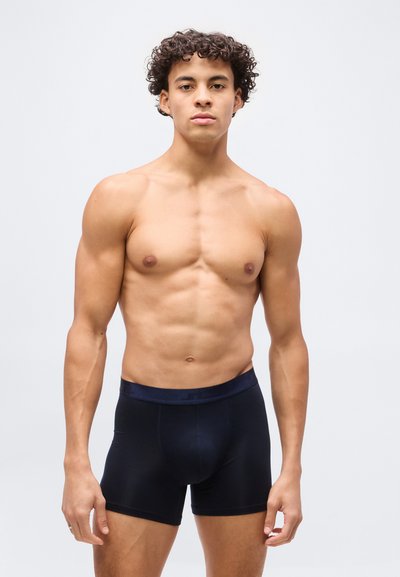 Νέος άνδρας με σγουρά μαλλιά στέκεται μπροστά, φοράει μαύρα εφαρμοστά μποξεράκια σε απλό φωτεινό φόντο.