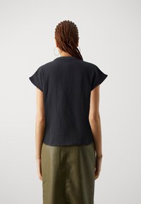 Donna con capelli intrecciati, che indossa orecchini a cerchio, una maglietta nera a maniche corte e una gonna verde oliva, in piedi con le spalle alla fotocamera.