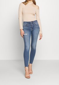 Vaahto beige ribattu pitkähihaine paita, jossa on olkapäältä avonainen muotoilu. Mallilla on päällään vaaleansiniset kapeat farkut, joissa on kulutusta polvessa.