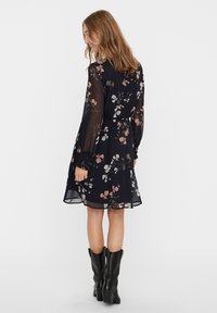 Robe noire à imprimé floral avec une taille cintrée, de longues manches transparentes et une longueur aux genoux. Présente des motifs floraux roses et blancs. Assortie avec des bottes noires.