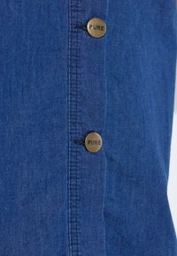 Blauwe denimstof met verticale stiksels en twee koperen knopen gegraveerd met het woord "PURE", uitgelijnd in een kolom.