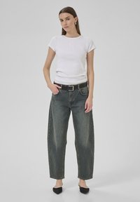 Balts ribēts T-krekls ar īsām piedurknēm un reljefu logo, savienots ar augstām, brīvām, tumša denima džinsām un melnu jostu.