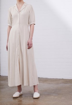 Robe boutonnée en lin beige avec manches courtes bouffantes et jupe longueur genou, associée à des espadrilles blanches sur un sol texturé.