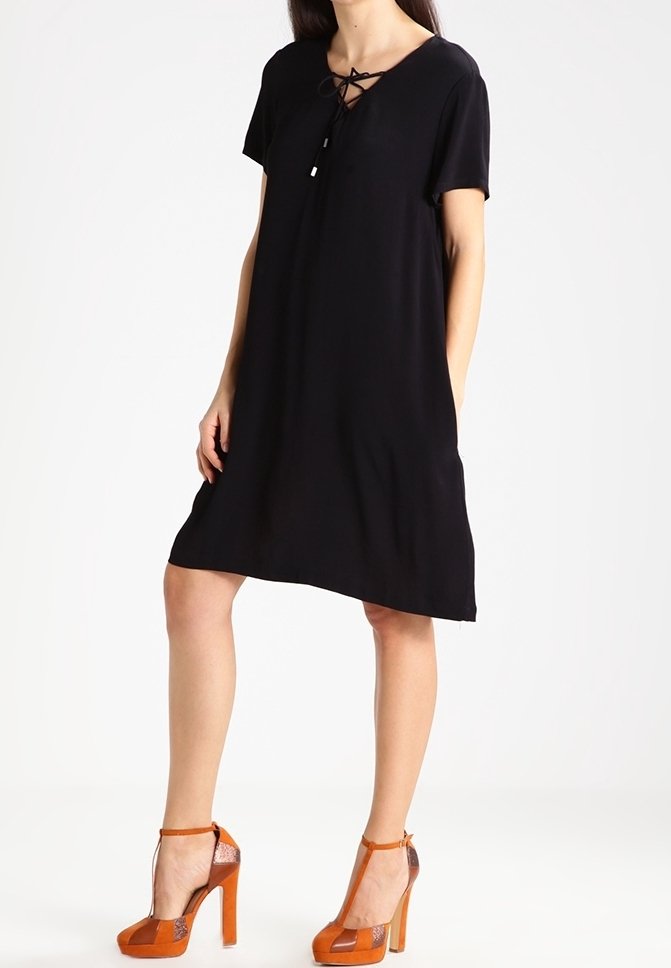 Robe courte noire à manches courtes avec un décolleté en V et un détail de lien en dentelle. Portée avec des sandales à talons orange ornées d'un accent transparent.