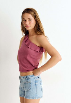 Junge Frau mit langen Haaren, die ein ärmelloses rosa Rüschentop und hellblaue Jeansshorts trägt, steht mit den Händen in der Hüfte.