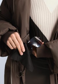 Čierna obdĺžniková čistiaca utierka držená v ruke, vedľa kovového predmetu s tmavým prechodovým povrchom, umiestnená na hnedej vypchávanej bunde.