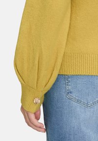 Senfgelbes Strickpullover mit ballonförmigen Ärmeln, ausgestattet mit gerippten Bündchen und einem floralem goldenen Knopfdetail. Kombiniert mit hellblauer Jeans.