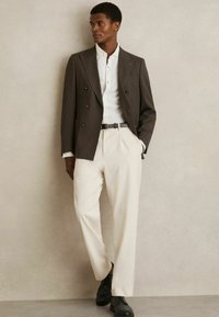 Dobbeltbrystet mørk blazer, hvid ribbet skjorte, cremefarvede plisserede bukser, sorte sko. Glatte teksturer, klassisk design, tætsiddende silhuet.