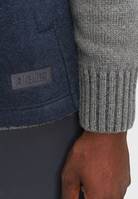 Giacca in misto lana blu navy con polsini a costine grigi, caratterizzata da una finitura texturizzata e da un discreto etichetta del brand sul lato sinistro.