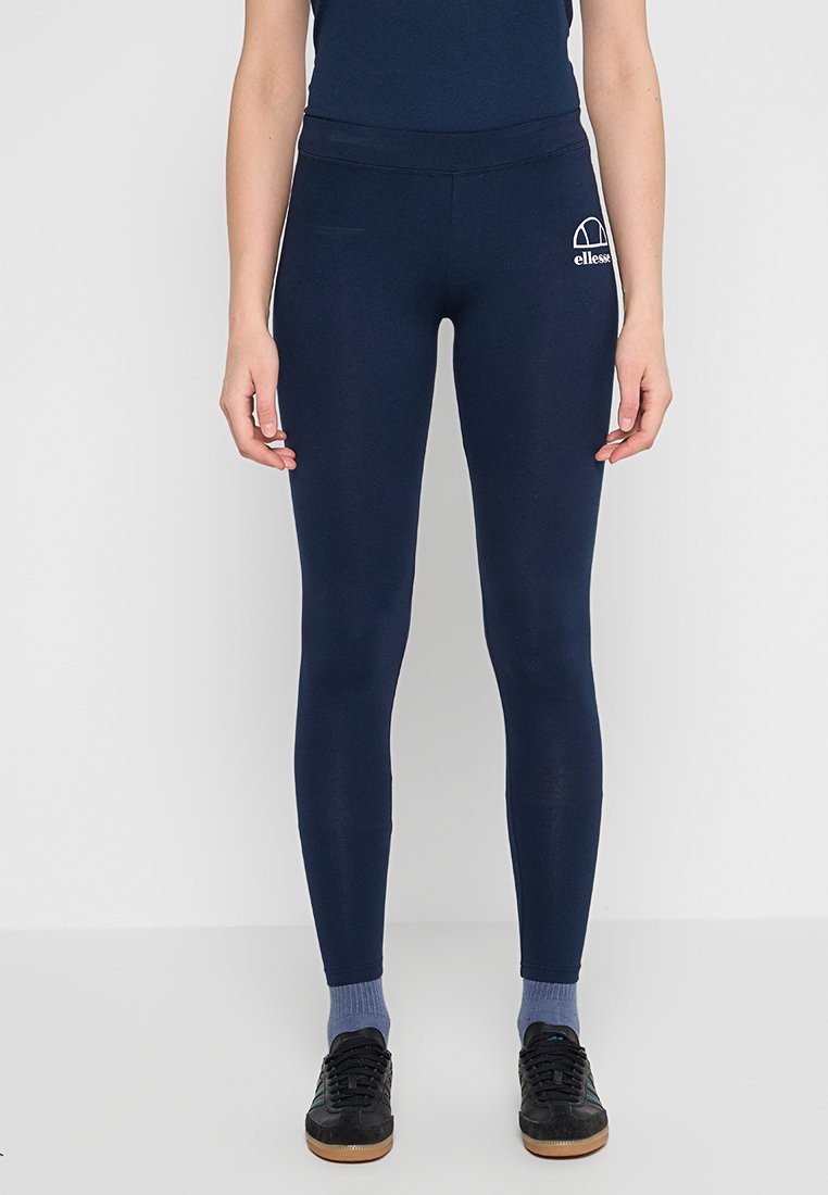 Donna che indossa leggings a vita alta blu scuro con il logo bianco Ellesse sulla coscia sinistra, calze blu e scarpe da ginnastica nere.