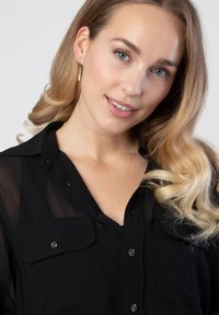 Zwarte sheer knopshirt met twee borstzakken, gedragen met een delicate gouden ketting. Het model heeft golvend blond haar en een neutrale achtergrond.