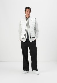 White padded vest with a quilted texture, black zipper, and logo detail, worn over a light gray shirt, paired with black pants and white sneakers.