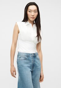 Γυναίκα με μακριά σκουρόχρωμα μαλλιά, φορώντας λευκό αμάνικο τοπ με γιακά και τζιν παντελόνι ψηλόμεσο σε ανοιχτό μπλε χρώμα, μπροστά από απλό φόντο.