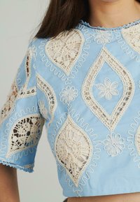 Blaues, gekürztes Hemd mit aufwendigen beigefarbenen Spitzenapplikationen in floralen und tropfenförmigen Mustern sowie gewellten Kanten an Ärmeln und Ausschnitt.