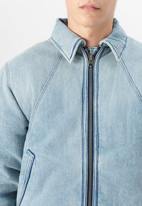 Veste en denim bleu clair avec un col montant, fermeture éclair à l'avant et manches raglan. Présente des coutures contrastantes et des poches latérales.