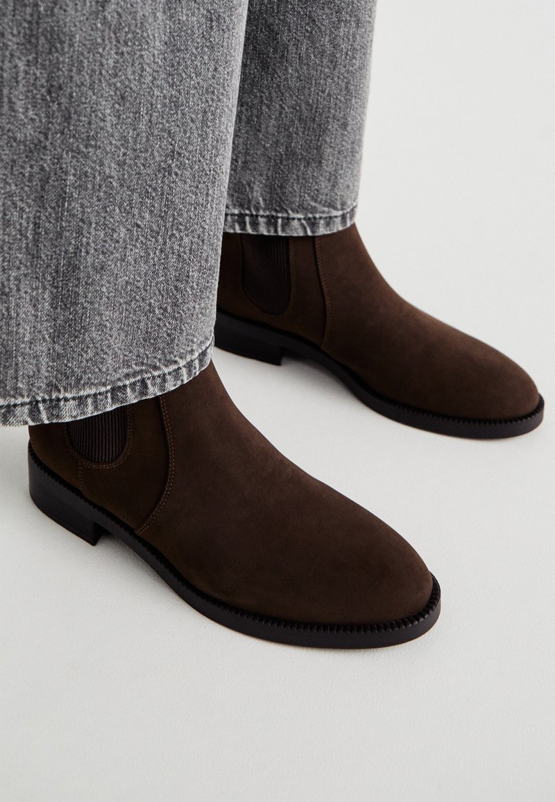 Bruine suède Chelsea boots met elastische zijpanelen, ronde neus en een gestructureerde zool. Gecombineerd met lichtgrijze losse spijkerbroek.
