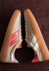 Paire de baskets en daim beige avec semelles en gomme, lacets blancs, une chaussure avec des rayures rouges, l'autre avec des rayures blanches, posées sur une surface texturée sombre.