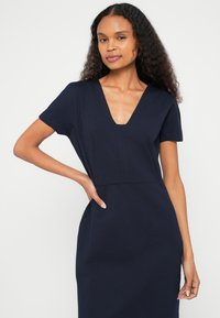 Tamsiai mėlyna suknelė su V formos iškirpte, trumpomis rankovėmis ir aptempta siluetu. Pagaminta iš lygaus audinio, su šoniniais kišenėmis ir minimaliais siūlėmis.