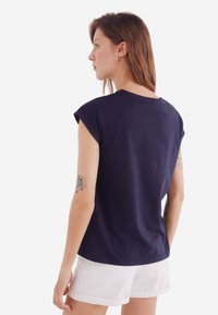 Marineblå bomulds t-shirt med rullede korte ærmer, afslappet pasform og rund halsudskæring, vist fra ryggen med lyse shorts.