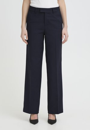 Femeie îmbrăcată cu pantaloni negri drepti, cu talie dreaptă, pantofi negri cu vârf ascuțit și un top alb, stând în picioare pe un fundal deschis simplu.