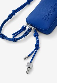 Modrá kožená crossbody taška s textúrovaným povrchom, obsahujúca zips, uzlový popruh a kovové akcenty.