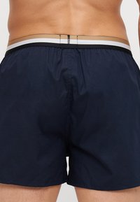 Navy swim trunks feature a contrasting waistband with stripes in gold, white, and black; made from lightweight, textured fabric.