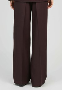 Vue arrière de pantalon large, brun foncé, ajusté, porté avec des chaussures noires, montrant des plis droits et une coupe décontractée du taille à l'ourlet.