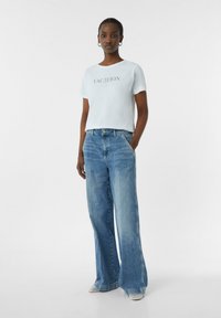 T-shirt en coton bleu clair avec le texte "VACANCES", associé à un jean large taille haute bleu clair. Détails texturés sur l'ourlet des chaussures.
