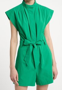 Combinaison courte en coton vert avec un col montant, des manches courtes, des plis et une taille ceinturée. Texture lisse avec une couleur unie et des détails minimaux.